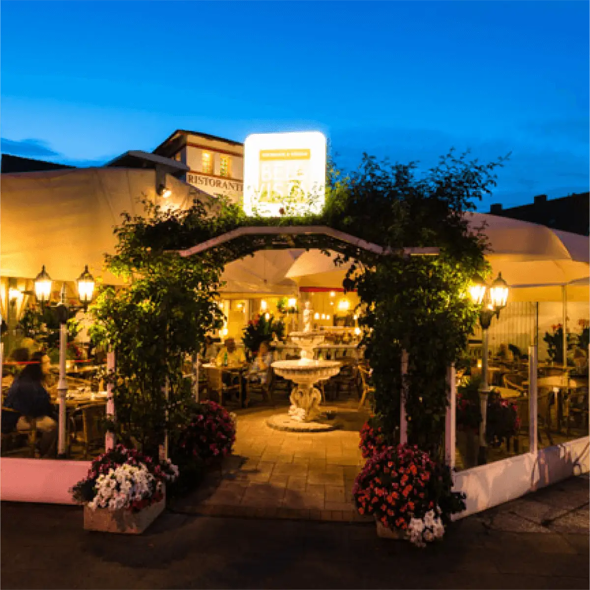 Eingangsbereich des Restaurants mit einem schönen Torbogen und vielen Blumen in Abenddämmerung.